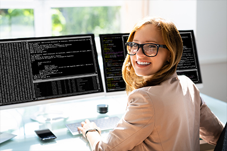 Focused female software developer working on code in a modern office setting, with lines of code visible on her dual monitors and a notepad beside her keyboard.